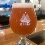 A tulip glass of amber beer with a foamy head, etched "MoMac Brewing Company" logo, on a wooden bar.