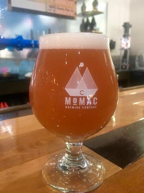 A tulip glass of amber beer with a foamy head, etched "MoMac Brewing Company" logo, on a wooden bar.