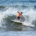 A shirtless man in navy swim trunks surfs a white board on a breaking wave, with other surfers in the background.