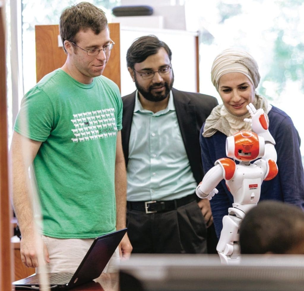 Three people watch a small white and orange humanoid robot while a laptop sits open on the table beside them.
