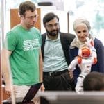 Three people watch a small white and orange humanoid robot while a laptop sits open on the table beside them.