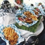Football-themed spread of burgers, fries, fried chicken bites and salads in checkered paper on turf, with navy helmets.