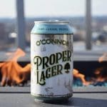 O'Connor Proper Lager beer can standing on a ledge in front of an outdoor fire pit with a city view.