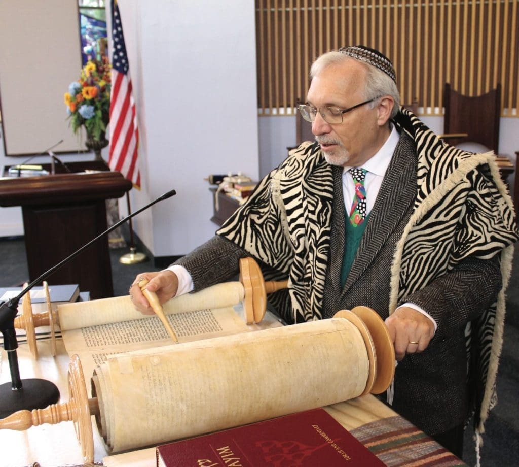A man in a zebra-print prayer shawl and kippah reads a Torah scroll with a yad pointer in a synagogue.