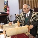 A man in a zebra-print prayer shawl and kippah reads a Torah scroll with a yad pointer in a synagogue.