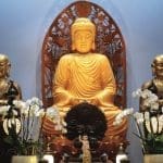 A golden seated Buddha statue flanked by two bronze monk figures, white orchids, and a carved wooden backdrop.