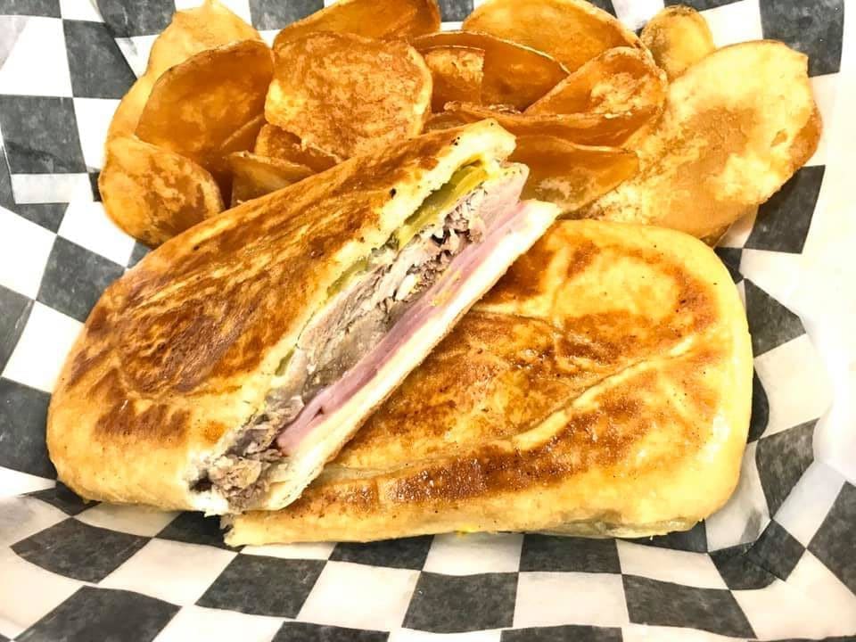 A pressed Cuban sandwich cut in half, showing pork, ham, pickles and cheese, with potato chips on checkered paper.