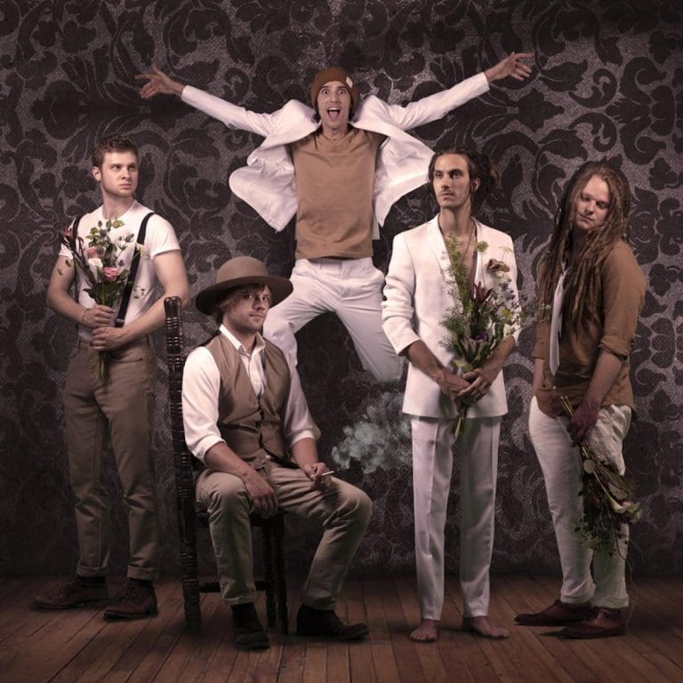 Five men in vintage-style clothes pose against dark damask wallpaper, one leaping midair and another sitting and smoking.
