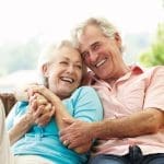 An older couple laughs together on a wicker sofa, his arms wrapped around her, her hands clasped with his.