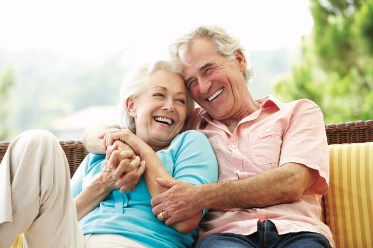 An older couple laughs together on a wicker sofa, his arms wrapped around her, her hands clasped with his.