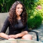 A smiling woman with long curly dark hair sits in a gray patio chair by a poolside garden.
