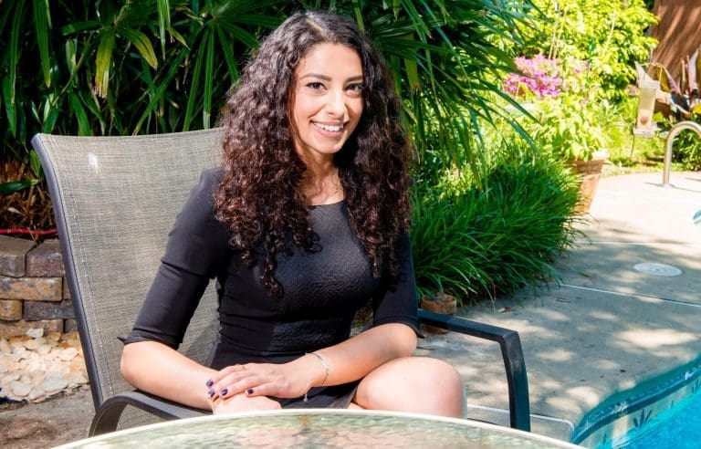 A smiling woman with long curly dark hair sits in a gray patio chair by a poolside garden.