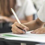 A student with long hair writes with a pen on paper at a desk in a classroom.