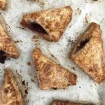 Golden triangular hand pies with sugar-crusted tops and dark berry filling on a parchment-lined baking sheet.