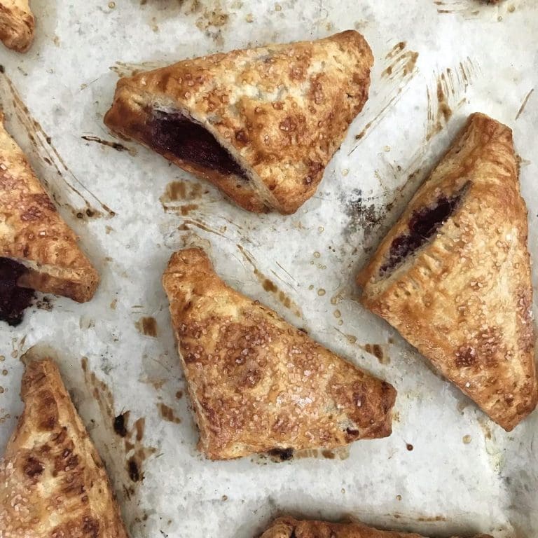 Golden triangular hand pies with sugar-crusted tops and dark berry filling on a parchment-lined baking sheet.
