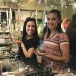 Two smiling women hold glass terrariums at a craft workshop, with sand, shells and plants on tables around them.