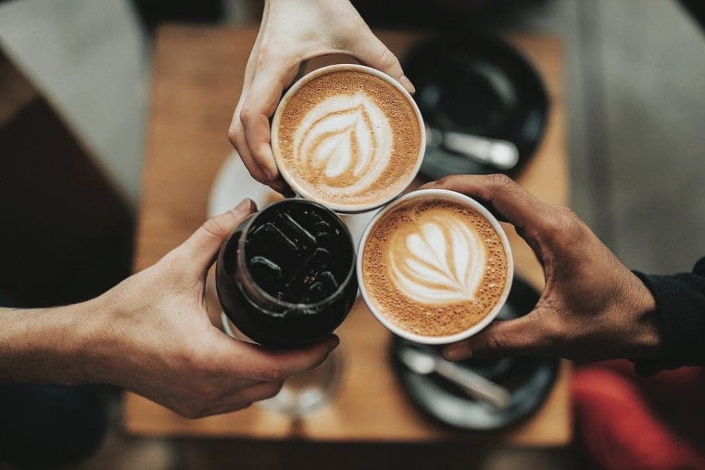 Three hands toast with two latte cups bearing leaf latte art and a dark iced coffee over a wooden table.