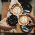 Three hands toast with two latte cups bearing leaf latte art and a dark iced coffee over a wooden table.