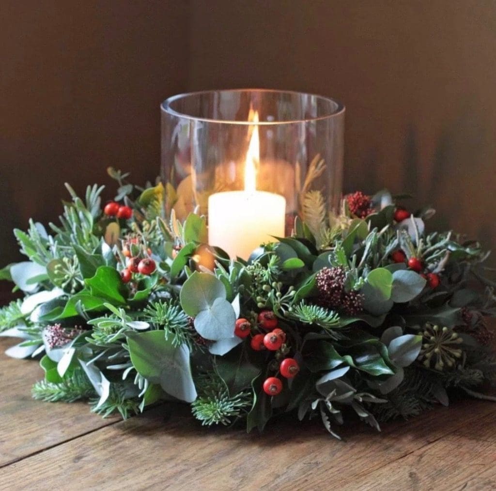 A glass hurricane candle surrounded by a holiday centerpiece of evergreens, eucalyptus, and red berries on a wooden table.