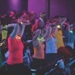 A group of people in bright tank tops wearing glow sticks cycles on stationary bikes in a dimly lit room.