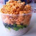 A clear plastic cup of poke bowl layered with white rice, seaweed salad, and orange spicy tuna with tofu cubes.