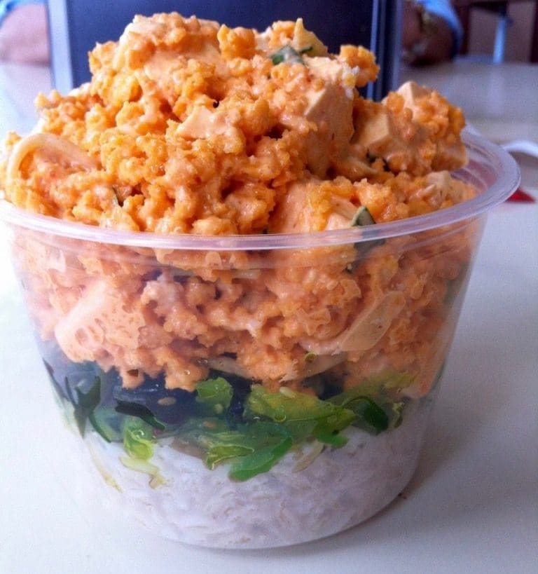 A clear plastic cup of poke bowl layered with white rice, seaweed salad, and orange spicy tuna with tofu cubes.