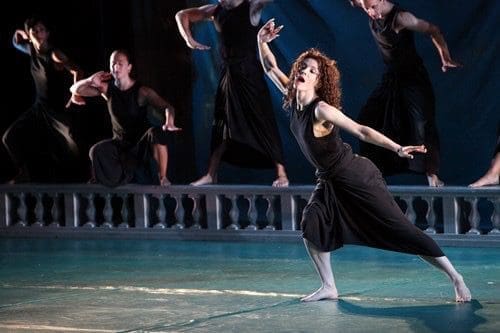 Barefoot dancers in black sleeveless costumes perform on a stage, with a curly-haired dancer in the foreground reaching one arm outward.