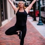 A woman in black athletic wear balances on one leg on a brick sidewalk, arms raised overhead in a city street.