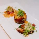 Three small plated dishes on a long white plate: a shredded slaw, a fried ball topped with microgreens, and seared octopus.