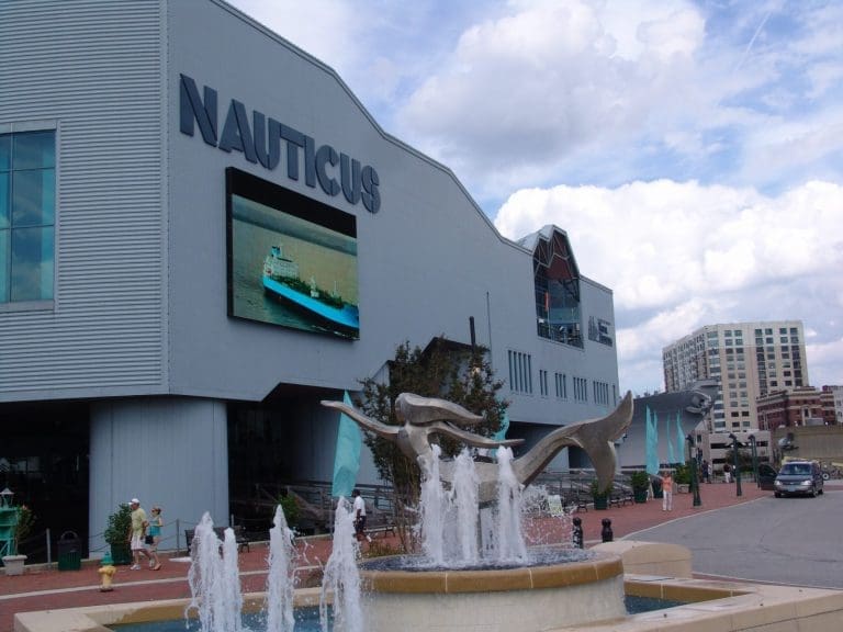 The Nauticus museum building in Norfolk, with a large screen showing a ship and a fountain with a sculpture out front.