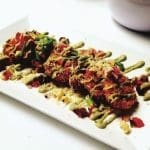 Crispy breaded fried dish on a white rectangular plate, drizzled with green sauce, topped with shaved cheese, scallions and corn.