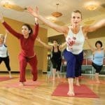 Five women in warrior pose with arms raised on red yoga mats in a bright wood-floored studio.