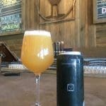 A stemmed glass of golden hazy beer beside a dark can labeled Commonwealth Brewing Co., with chalkboard beer menus behind.