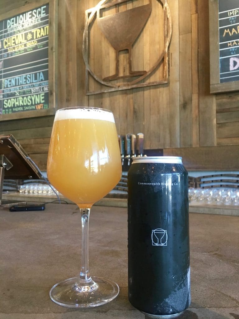 A stemmed glass of golden hazy beer beside a dark can labeled Commonwealth Brewing Co., with chalkboard beer menus behind.