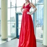 A woman in a long red velvet gown with a train, white gloves and a turban, posing in a glass-walled lobby.