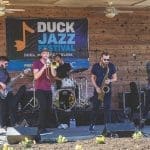 Six musicians perform on a wooden stage under a "Duck Jazz Festival" banner, with trombone, saxophone, trumpet, bass, and drums.