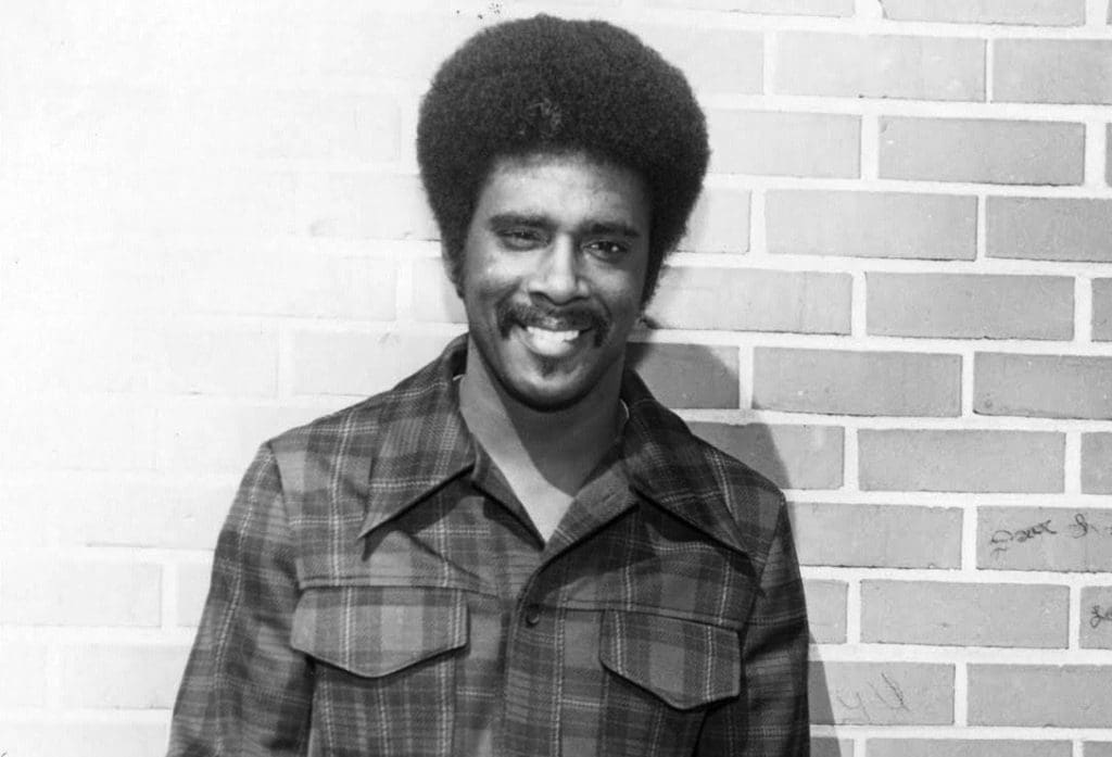 A smiling man with a full afro and mustache wears a plaid shirt, standing before a brick wall in black and white.