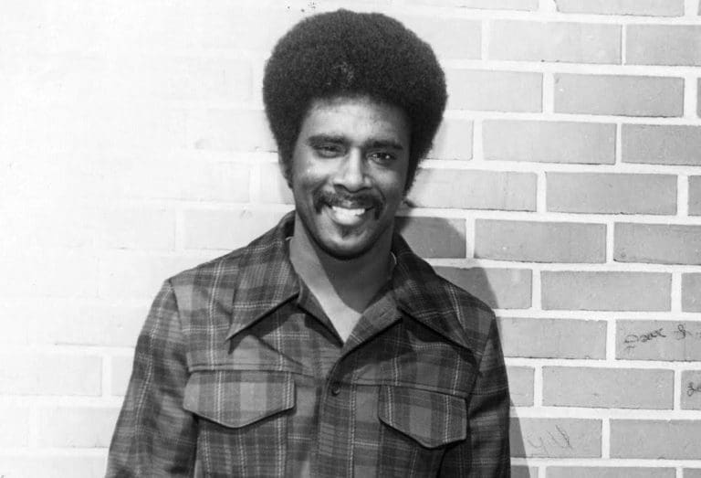 A smiling man with a full afro and mustache wears a plaid shirt, standing before a brick wall in black and white.