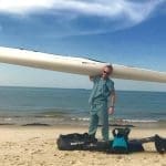A man in scrubs holds a white Epic kayak overhead on a sandy beach, with gear bags at his feet.