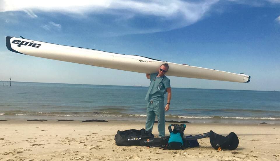 A man in scrubs holds a white Epic kayak overhead on a sandy beach, with gear bags at his feet.
