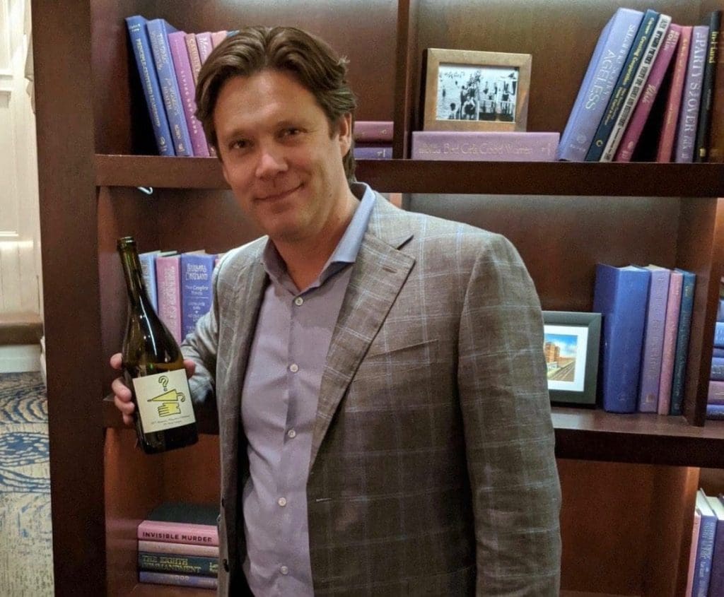 A smiling man in a gray plaid blazer holds a bottle of wine in front of a wooden bookshelf.