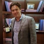 A smiling man in a gray plaid blazer holds a bottle of wine in front of a wooden bookshelf.