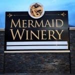 Monument sign reading "Mermaid Winery" in gold letters with a gold mermaid emblem, set on a stone-clad base at dusk.