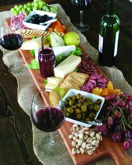 A long wooden charcuterie board on burlap holds cheeses, crackers, olives, pistachios, grapes, fruit, and two glasses of red wine.
