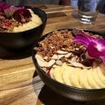 Two black bowls of granola-topped acai bowls with sliced banana, almond slices, and purple orchid garnish on a wooden table.