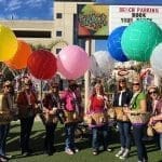 People in aviator caps hold wicker baskets beneath colorful balloons in front of The Shack on 8th sign.