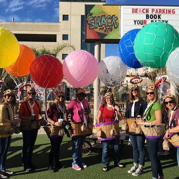 People in aviator caps hold wicker baskets beneath colorful balloons in front of The Shack on 8th sign.