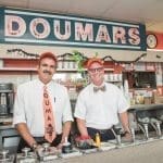 Two smiling men in white shirts and orange caps stand behind the counter of Doumars, under its red neon sign.