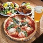 Margherita pizza with basil and tomato, fried chicken with celery, Greek salad, and a glass of beer on a wooden table.