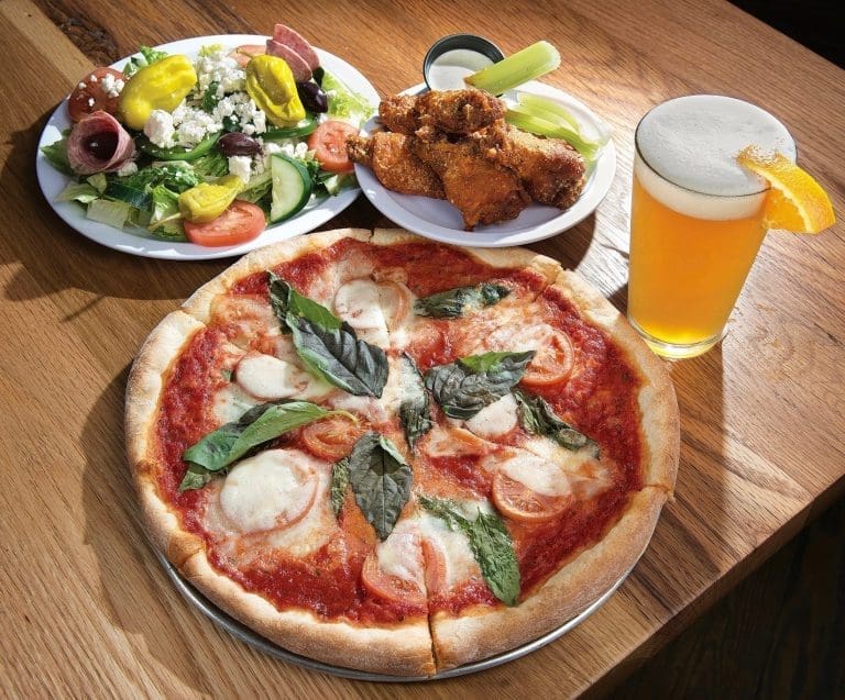 Margherita pizza with basil and tomato, fried chicken with celery, Greek salad, and a glass of beer on a wooden table.